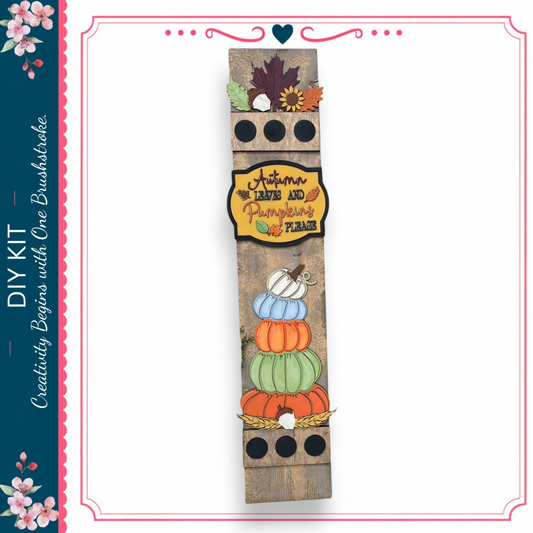 a wooden sign with a decorative design, including a stack of pumpkins and autumn leaves. The sign is surrounded by a border and has a heart-shaped decoration at the top.