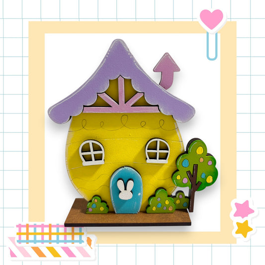 A colorful, whimsical toy house with a purple roof, white windows, and a blue door, set against a grid background with a heart and stars.