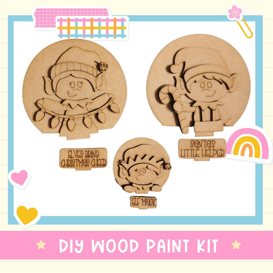 The image is a DIY wood paint kit featuring three wooden shapes resembling characters, including a Christmas elf, a little helper, and a magic elf.