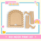 The image is a DIY wood paint kit, featuring a wooden frame with a scalloped edge and two wooden pieces, one of which is a wooden boot. The kit is presented against a light blue background with a pink border.