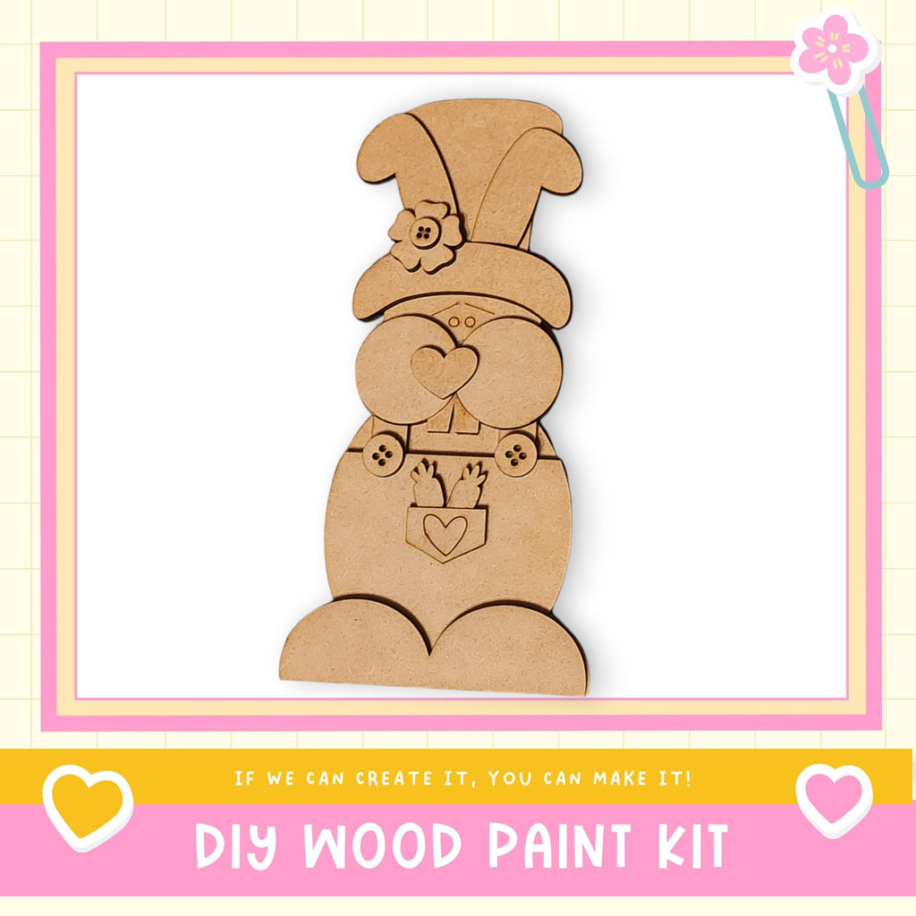 a DIY wood paint kit, with a wooden gnome figure as the main focus. The gnome is wearing a hat with a flower and has a heart-shaped bow around its neck. The kit appears to be made of wood and is designed for creating wooden figures.