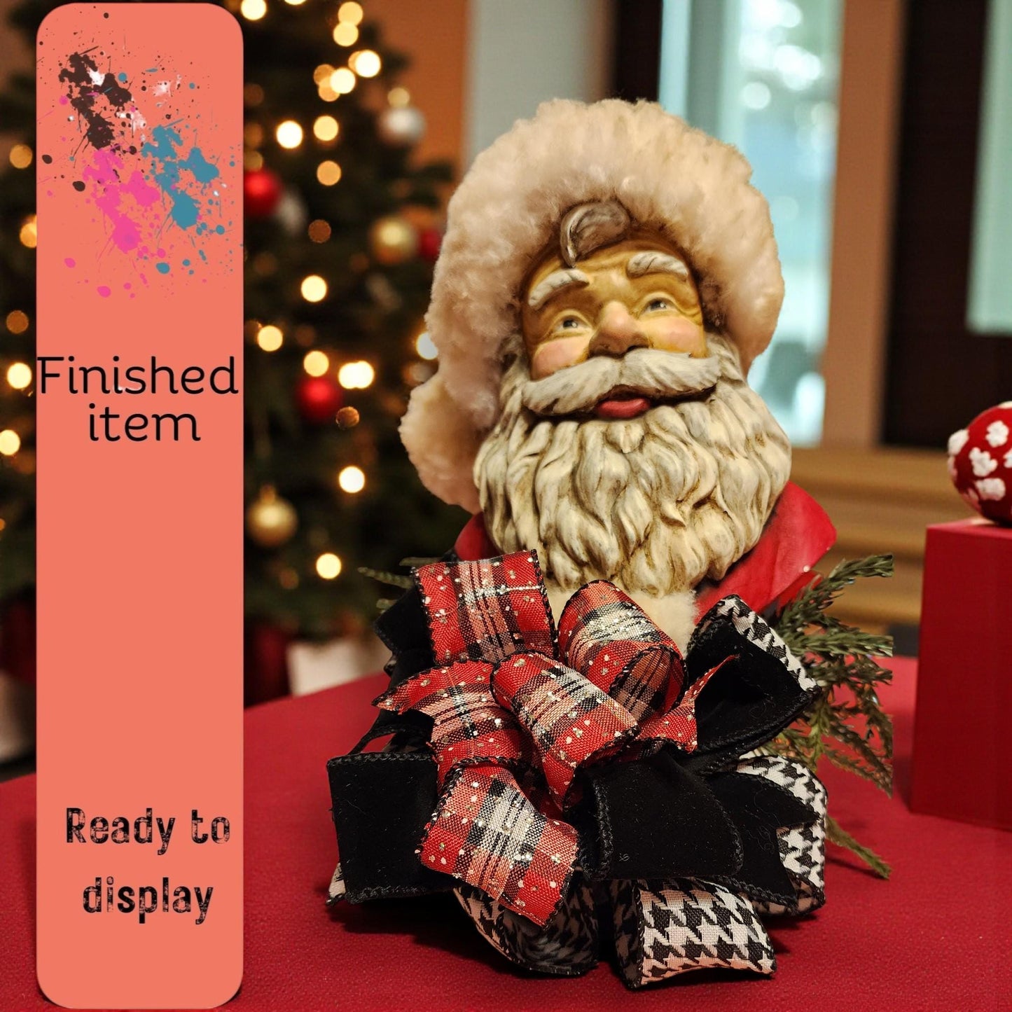 A Santa Claus figurine with a red and black plaid bow sits on a table, with a Christmas tree and other holiday decorations visible in the background.