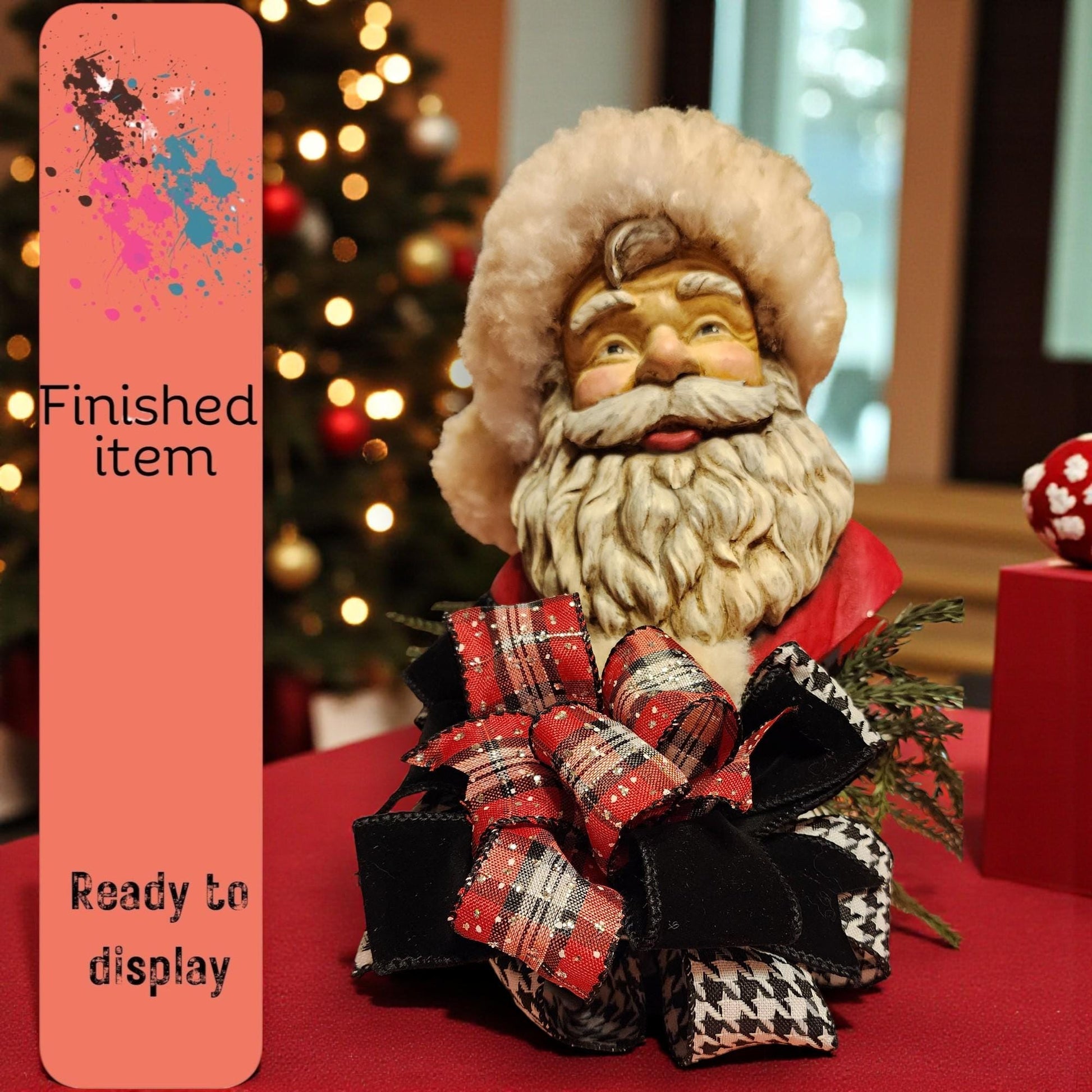 A Santa Claus figurine with a red and black plaid bow sits on a table, with a Christmas tree and other holiday decorations visible in the background.