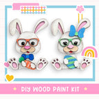 two cute wooden rabbits wearing glasses and holding Easter eggs, with a DIY wood paint kit labeled "DIY WOOD PAINT KIT" in the background.