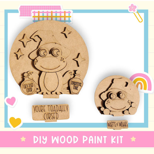 a DIY wood paint kit with a cute frog design. The kit includes a larger frog-shaped piece and a smaller frog-shaped piece, both with a whimsical and playful appearance. The kit also includes a sign that reads "You're Toadally Cursed" and a smaller sign that says "Warts & Wishes". The kit is presented in a colorful and playful manner, making it an appealing and fun project for children and adults alike.