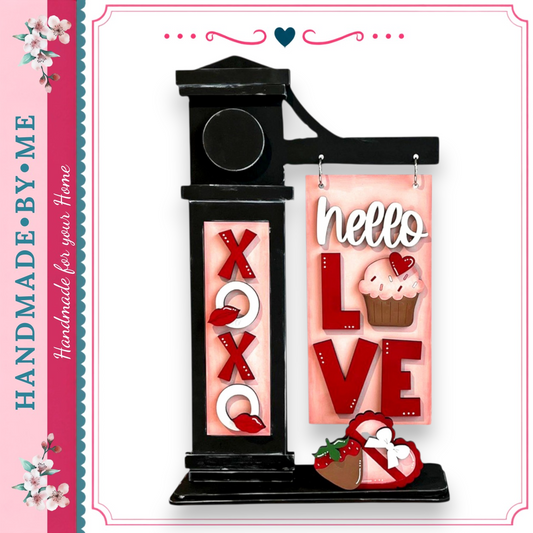 A handmade sign with the words "Hello Love" displayed on it, along with a cupcake and a heart. The sign is black and features decorative elements such as the word "XO" and the word "LOVE".