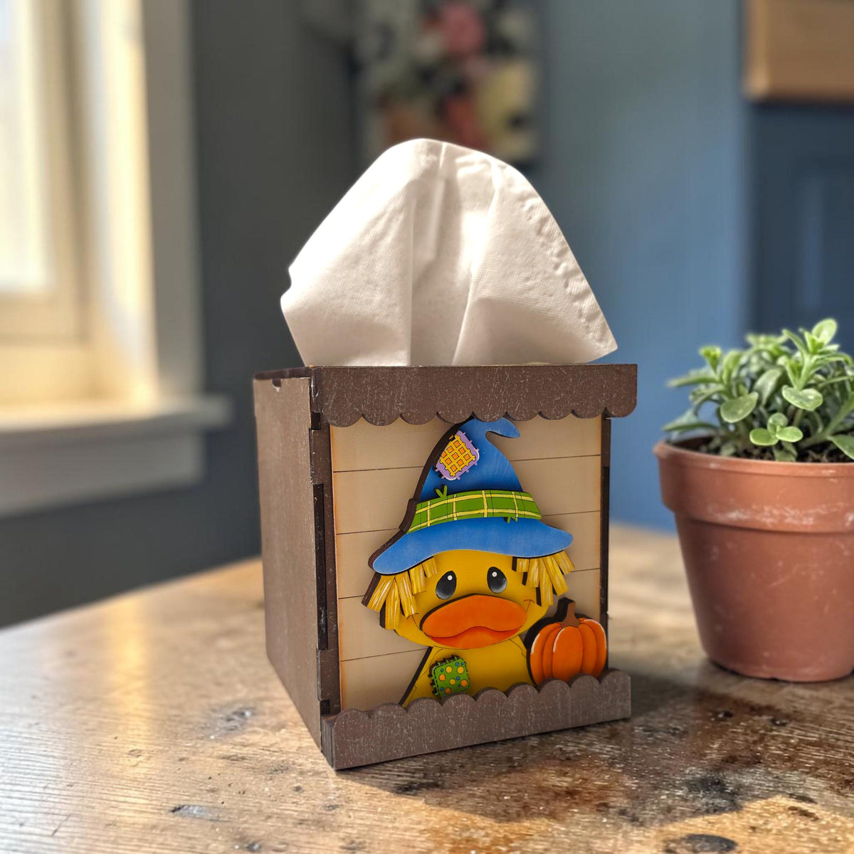 A wooden tissue box with a cartoon duck wearing a witch's hat and holding a pumpkin, placed on a wooden table next to a potted plant.