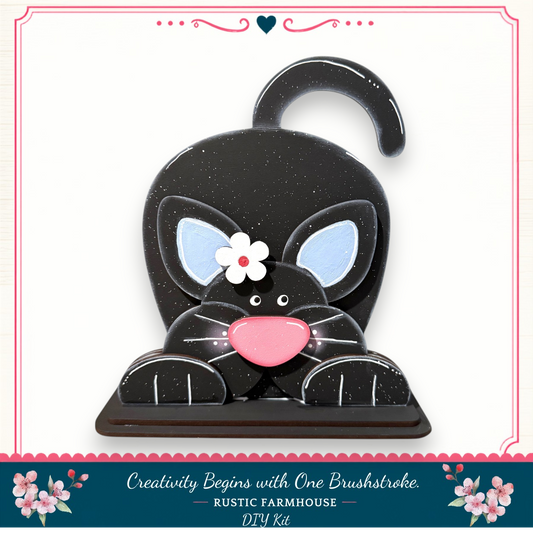A black cat-shaped object with a pink nose and a white flower on its face, sitting on a wooden base.