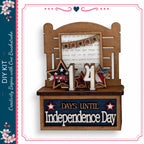 The image is a wooden display with a sign that reads "DAYS UNTIL Independence Day" and a banner with the number "14" and stars. The display is set against a white background, and there is a text box with a message that reads "I can see by the dawn, when the stars and stripes are through the perils, and the remnants were so clearly streaming."