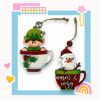 two Christmas ornaments, one of a elf and one of a snowman, both sitting inside a cup. The elf is wearing a green hat and the snowman is wearing a red hat. The cup has the words "Warm & Cozy" written on it. The background of the image is a light blue color with a pink and white checkered pattern.