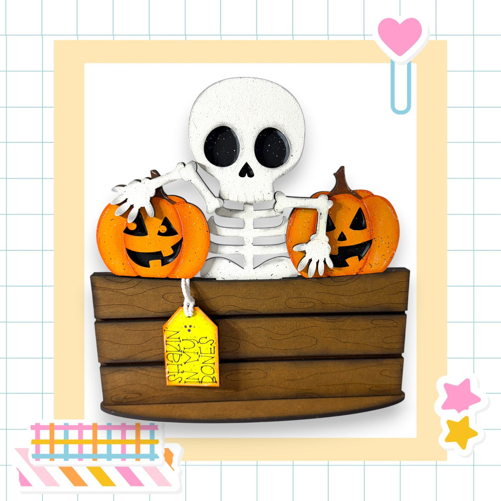 A skeleton holding two carved pumpkins, sitting in a wooden crate, with a yellow tag attached to the crate.