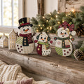 A festive scene with three colorful snowman decorations on a wooden shelf, surrounded by pine cones and greenery, with a window and string lights in the background.