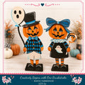 two wooden Halloween decorations, a jack-o-lantern and a ghost, standing on a wooden surface. The decorations are dressed in black and blue outfits, and they are surrounded by pumpkins and other Halloween-themed decorations.