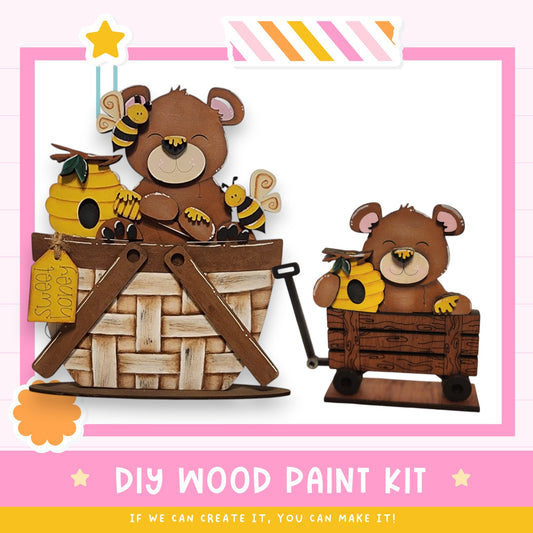 two wooden bears sitting in a basket, with one bear holding a honey pot. The bears are surrounded by various decorative elements, including a bee and a star. The image also includes text that reads "DIY WOOD PAINT KIT" and "IF WE CAN CREATE IT, YOU CAN MAKE IT!"