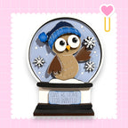 A snow globe containing a cartoon owl wearing a blue hat and holding snowflakes, with the text "OWL BE HOME FOR WINTER" on the base.