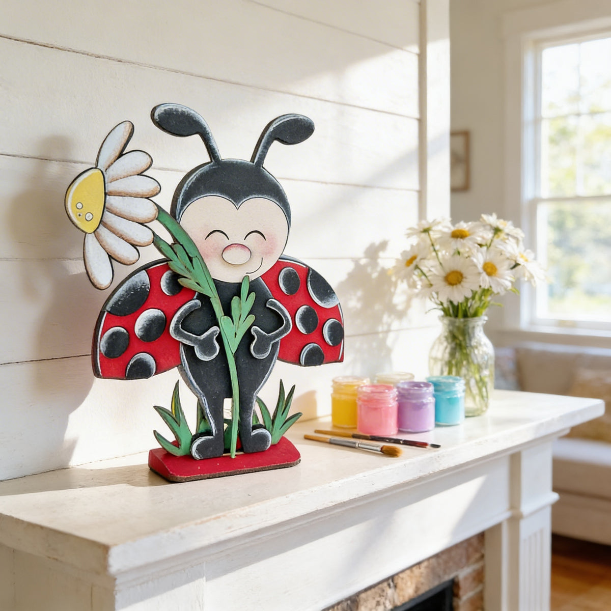 A wooden ladybug figurine holding a daisy flower is displayed on a white shelf, accompanied by a vase of white daisies and art supplies.