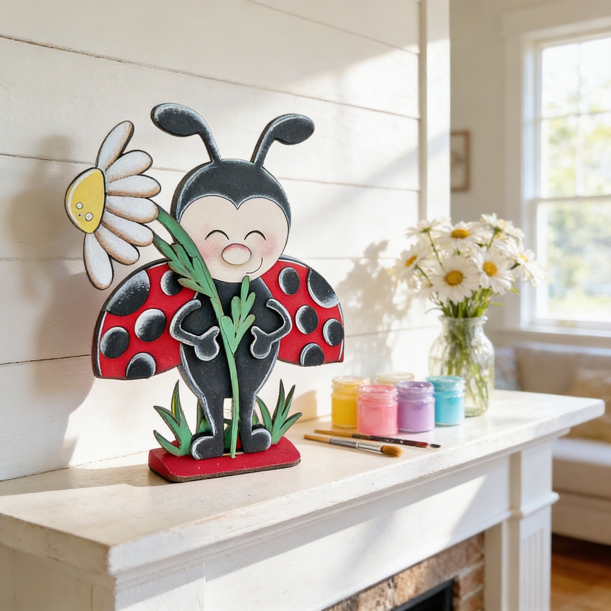 A wooden ladybug figurine holding a daisy flower is displayed on a white shelf, accompanied by a vase of white daisies and art supplies.