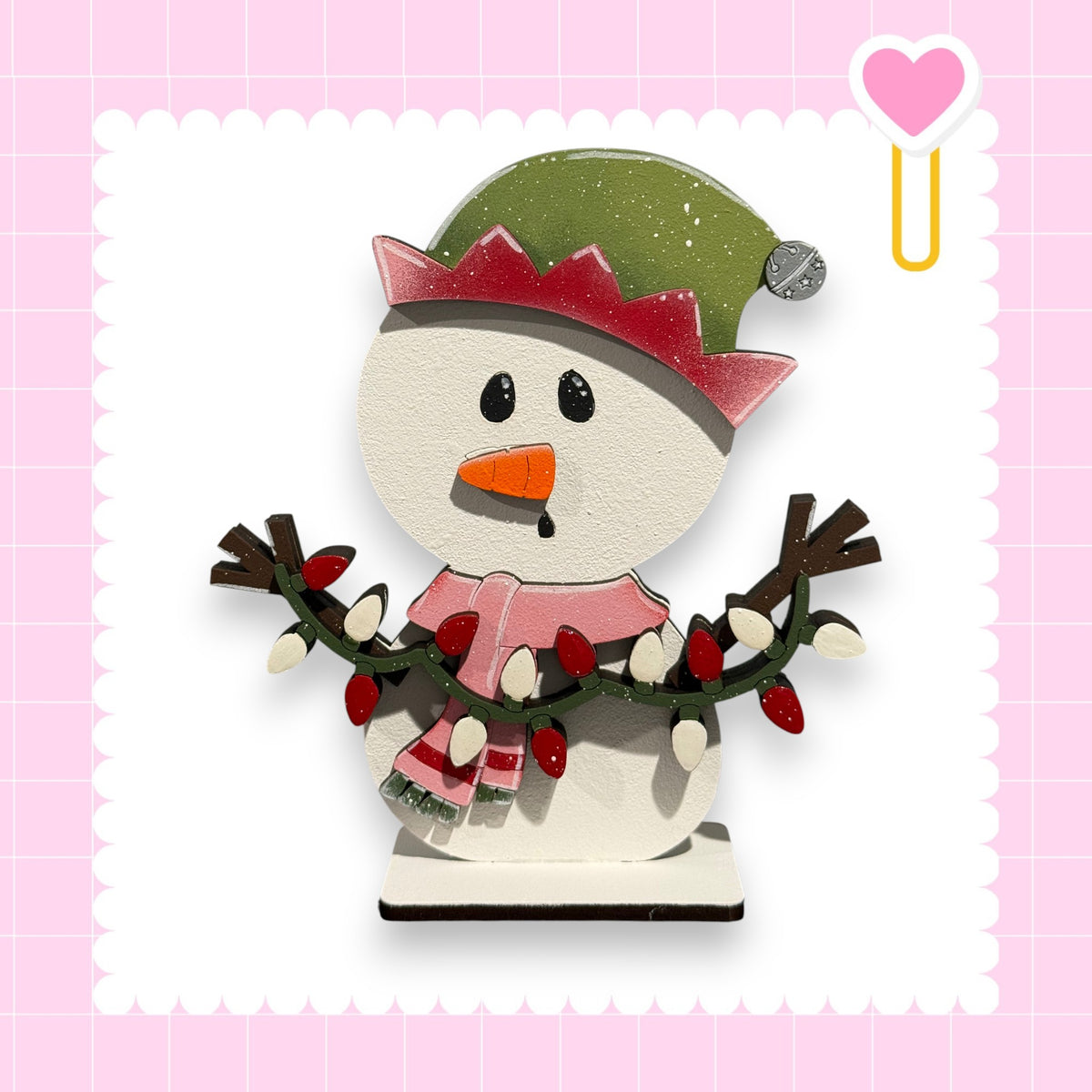 A wooden snowman figure with a green hat, red scarf, and a heart-shaped ornament on its head, set against a pink background with a scalloped edge.