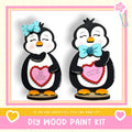 two wooden penguin figurines, each holding a heart-shaped sign with a Valentine's Day message. The penguins are wearing bows and appear to be made of wood. The image also includes text that says "DIY WOOD PAINT KIT" and "IF WE CAN CREATE IT, YOU CAN MAKE IT!"