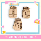 The image is a DIY wood paint kit featuring three wooden ornaments, including a mason jar, a snowman, and a rainbow.