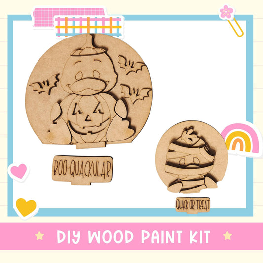 The image is a DIY wood paint kit featuring two wooden shapes, one resembling a pumpkin and the other a face, along with the text "BOO-QUACKULAR" and "QUACK OR TREAT".