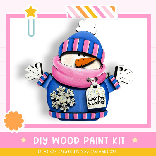 A wooden snowman figure wearing a pink and blue striped sweater and a blue hat with a white stripe, holding a carrot and wearing a "sweater weather" tag. The image is framed by a pink border with a yellow star and a white cloud.
