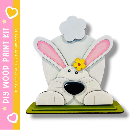 A wooden bunny sculpture with pink ears and a yellow flower on its head, sitting on a green base.