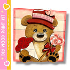 A wooden teddy bear holding a heart-shaped lollipop and a red bow, set against a pink background.