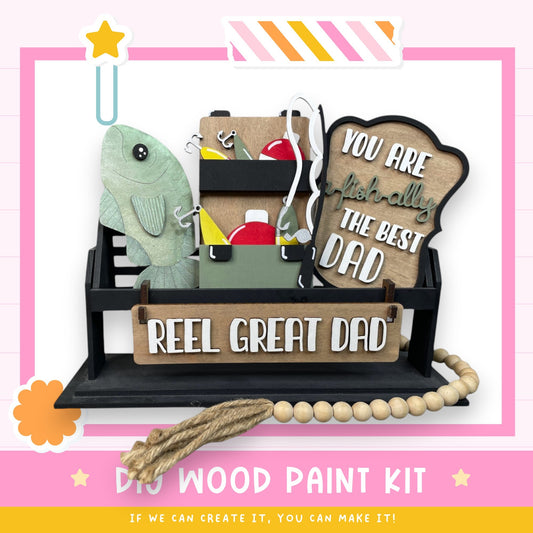 The image is a DIY wood paint kit with a wooden sign that says "REEL GREAT DAD" and a fish-shaped decoration. The kit appears to be made of wood and includes various tools and materials for painting.