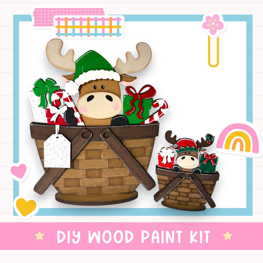 A wooden paint kit featuring a moose and a smaller moose in a basket, surrounded by various holiday-themed items such as presents and a bow.