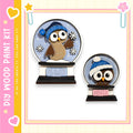 two snowglobe ornaments, each depicting an owl wearing a winter hat. The snowglobe on the left is larger and has a blue hat, while the one on the right is smaller and has a white hat. The snowglobes are set against a pink and yellow background, and the text "My Wood Paint Kit" is visible, suggesting that these ornaments are part of a kit for creating wooden decorations.