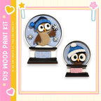 two snowglobe ornaments, each depicting an owl wearing a winter hat. The snowglobe on the left is larger and has a blue hat, while the one on the right is smaller and has a white hat. The snowglobes are set against a pink and yellow background, and the text "My Wood Paint Kit" is visible, suggesting that these ornaments are part of a kit for creating wooden decorations.
