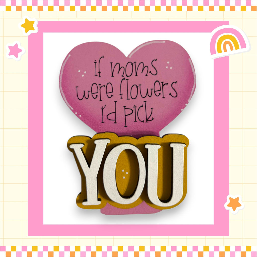 a heart-shaped sign with the text "If moms were flowers I'd pick you" written on it, along with the word "YOU" in a larger font. The sign is surrounded by a pink border and is set against a background with a checkered pattern.