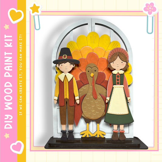 A wooden paint kit depicting a boy in a red and yellow outfit and a girl in a green and white outfit standing in front of a turkey, with a star and heart decoration on the left side.