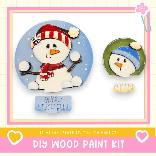 two snowman decorations, one larger and one smaller, both wearing winter hats and scarves. The larger snowman is wearing a red and white striped hat and a red scarf, while the smaller snowman is wearing a blue hat and a green scarf. The image also includes text that says "If we can create it, you can make it!" and "DIY WOOD PAINT KIT", suggesting that these snowman decorations can be easily created using a DIY wood paint kit.