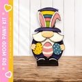 A wooden gnome figurine holding Easter eggs, wearing a colorful hat, stands on a wooden surface against a white background.