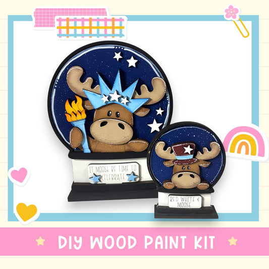 a DIY wood paint kit for creating a snow globe with a moose design. The kit includes a snow globe base and a moose figurine, along with a sign that reads "It's Moose Be Kind to Celebrate".