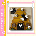 A wooden painting kit featuring a beehive with two bees and white flowers, along with a black door and windows, set against a yellow and pink background.