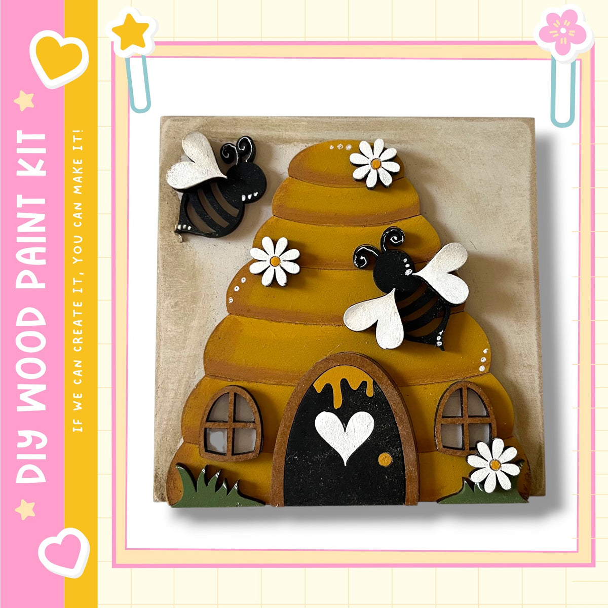 A wooden painting kit featuring a beehive with two bees and white flowers, along with a black door and windows, set against a yellow and pink background.