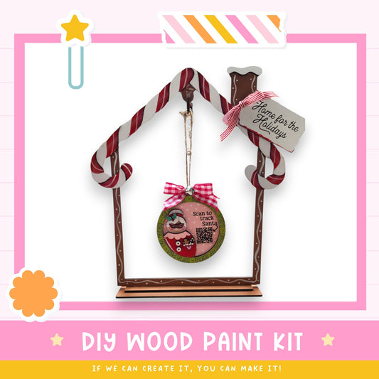 a DIY wood paint kit for creating a wooden ornament in the shape of a house. The kit includes a wooden frame, a candy cane decoration, and a sign that says "Home for the Holidays". The kit is presented in a pink and white color scheme, with a star decoration on the left side. The text "DIY WOOD PAINT KIT" is displayed at the bottom of the image, along with the phrase "If we can create it, you can make it!"