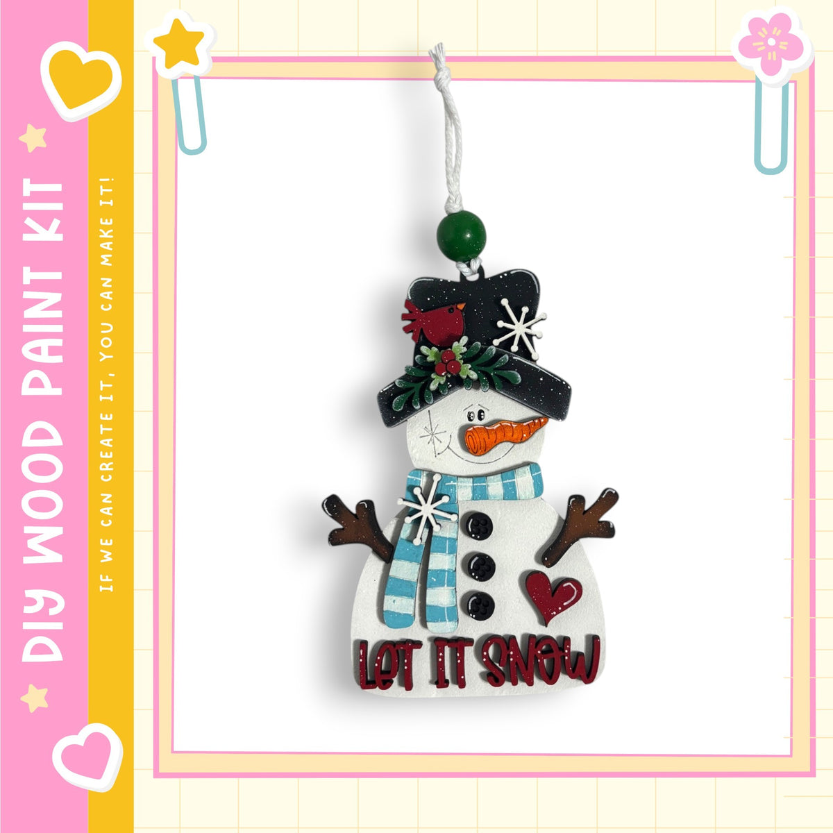 A snowman ornament with a carrot nose, scarf, and hat, hanging from a string. The snowman has a heart-shaped button on its chest and the text "LET IT SNOW" written on it.