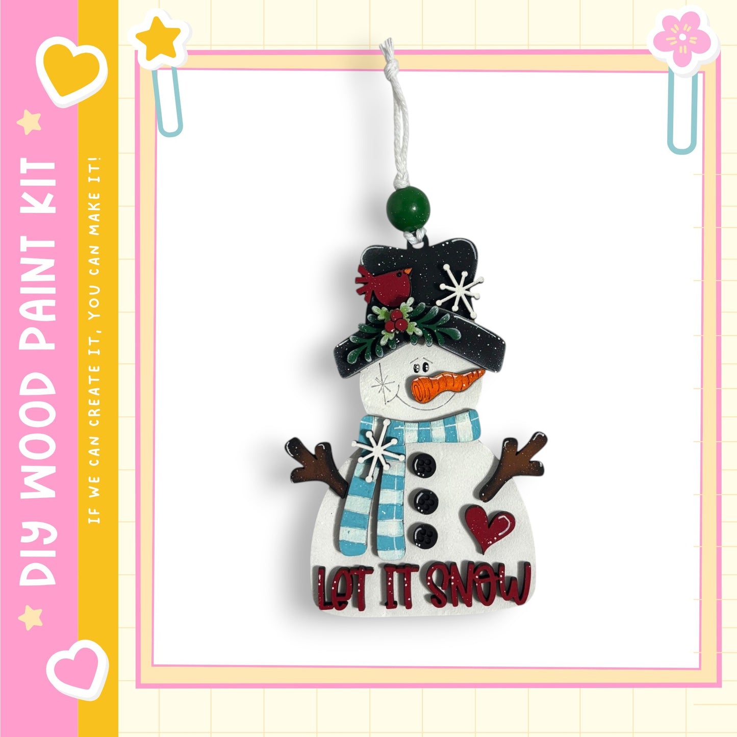A snowman ornament with a carrot nose, scarf, and hat, hanging from a string. The snowman has a heart-shaped button on its chest and the text "LET IT SNOW" written on it.