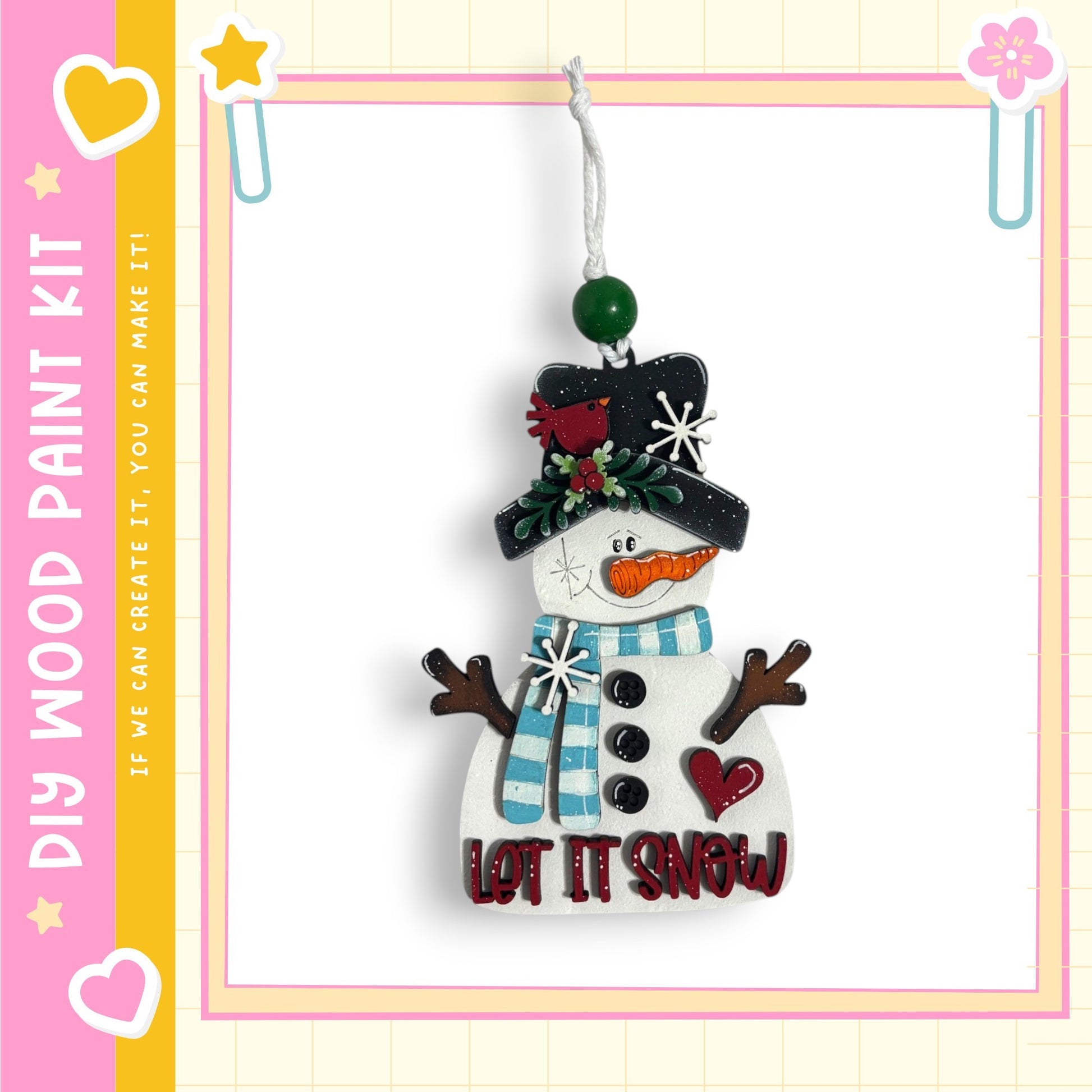 A snowman ornament with a carrot nose, scarf, and hat, hanging from a string. The snowman has a heart-shaped button on its chest and the text "LET IT SNOW" written on it.