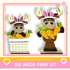 two wooden moose figurines, each holding a basket of carrots. The moose are wearing antlers and have pink ears. The image also includes text that says "DIY WOOD PAINT KIT" and "If we can create it, you can make it!"