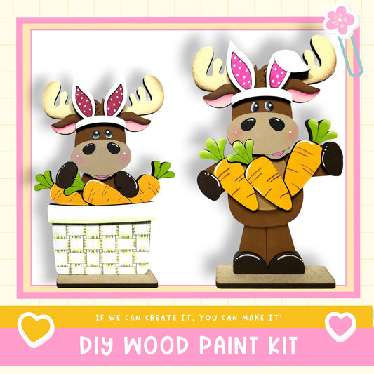 two wooden moose figurines, each holding a basket of carrots. The moose are wearing antlers and have pink ears. The image also includes text that says "DIY WOOD PAINT KIT" and "If we can create it, you can make it!"