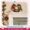 A handmade wooden decoration featuring a moose sitting on a shelf above a door, with Christmas-themed elements such as a wreath, a Santa card, and a postcard.