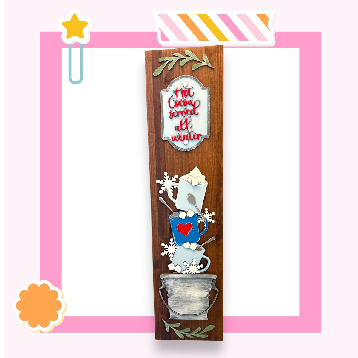 A wooden sign with a decorative design, featuring a heart-shaped mug, snowflakes, and a star, against a pink background.
