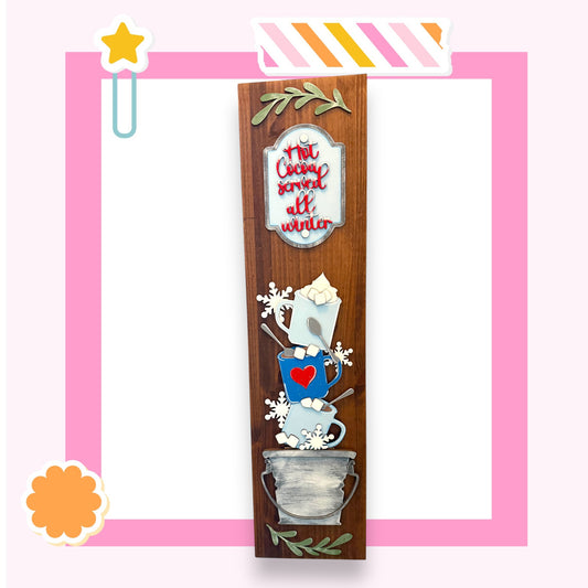 A wooden sign with a decorative design, featuring a heart-shaped mug, snowflakes, and a star, against a pink background.