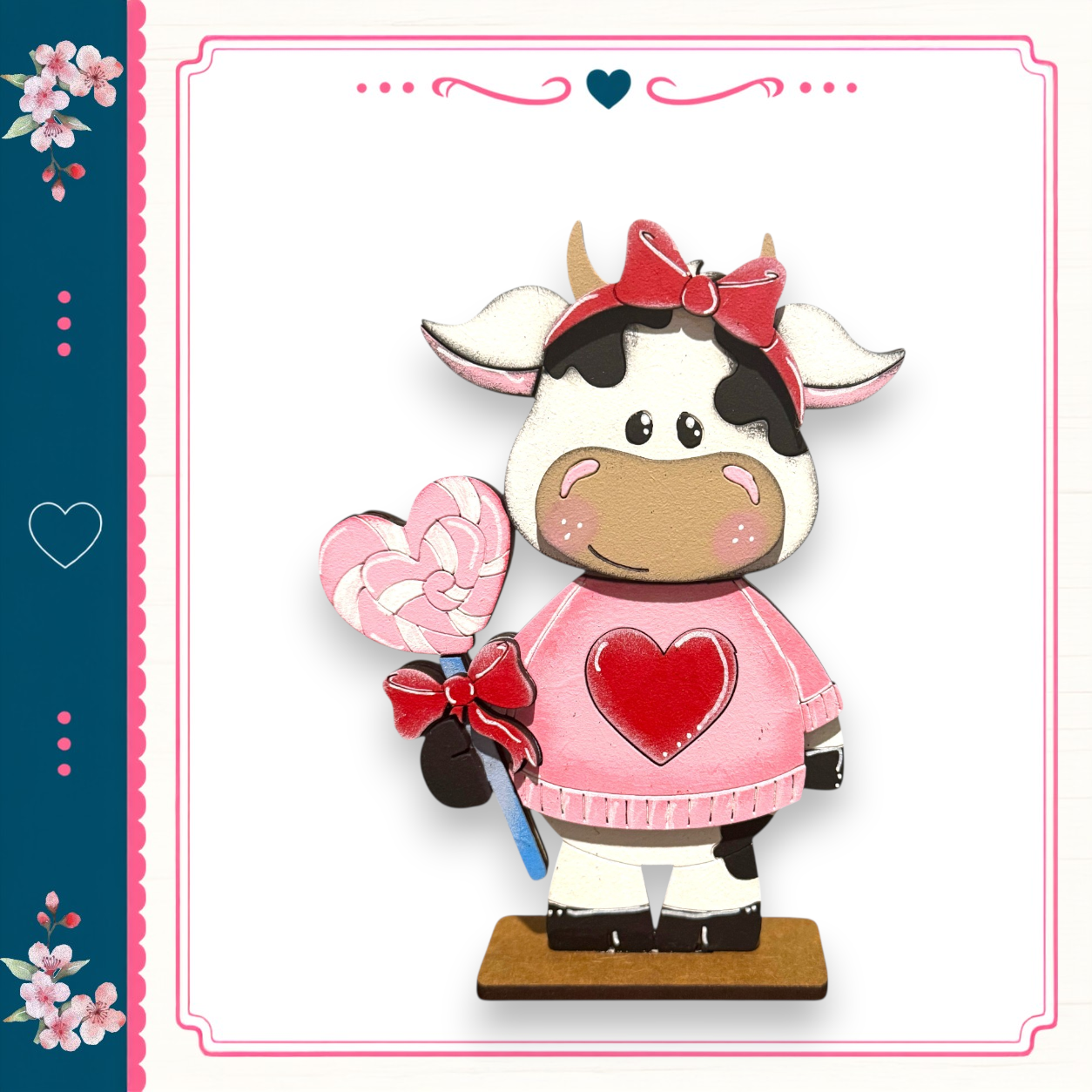 A wooden cow figurine wearing a pink sweater and holding a heart-shaped lollipop, standing on a wooden base.