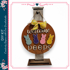a wooden sign with the words "Welcome Peeps" written on it, hanging on a wooden stand. The sign has four different colored Easter bunny shapes, including a blue bunny, a pink bunny, a purple bunny, and a yellow bunny. The sign is displayed against a white background, and there is a decorative border around the edges.